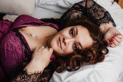 Woman in purple lingerie laying on bed smiling sweetly at camera. Boudoir in southern wisconsin. madison, WI boudoir photography